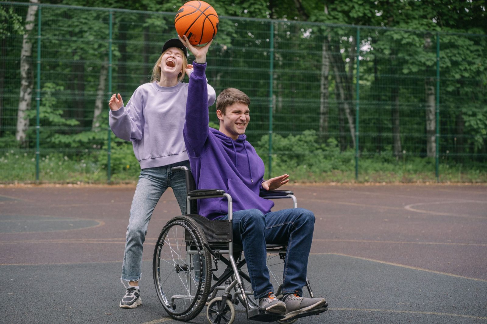 A young man in a wheelchair shoots a basketball on an outdoor court while a friend laughs behind him.