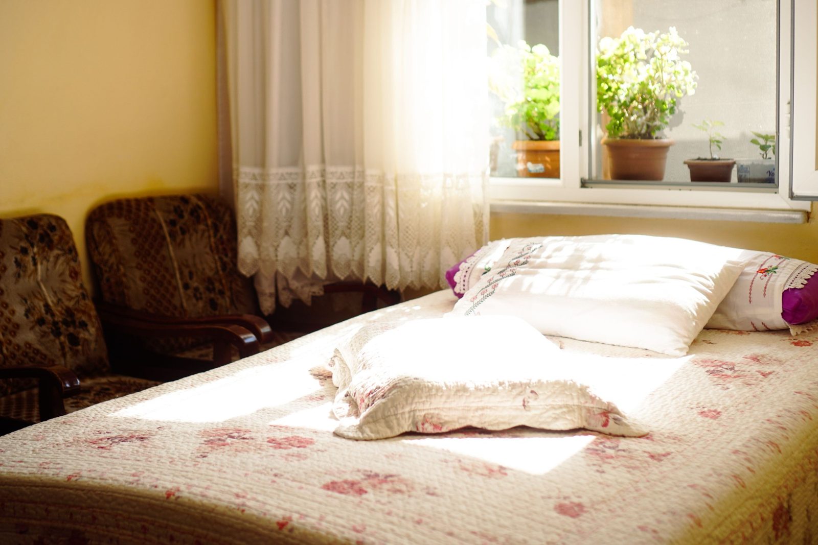 A sunlit bedroom with a floral quilt, lace curtains and plants on the windowsill.