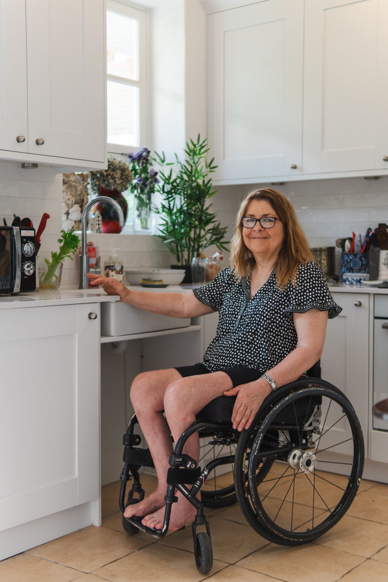 A woman using a wheelchair at her kitchen bench, one hand resting beside the tap.