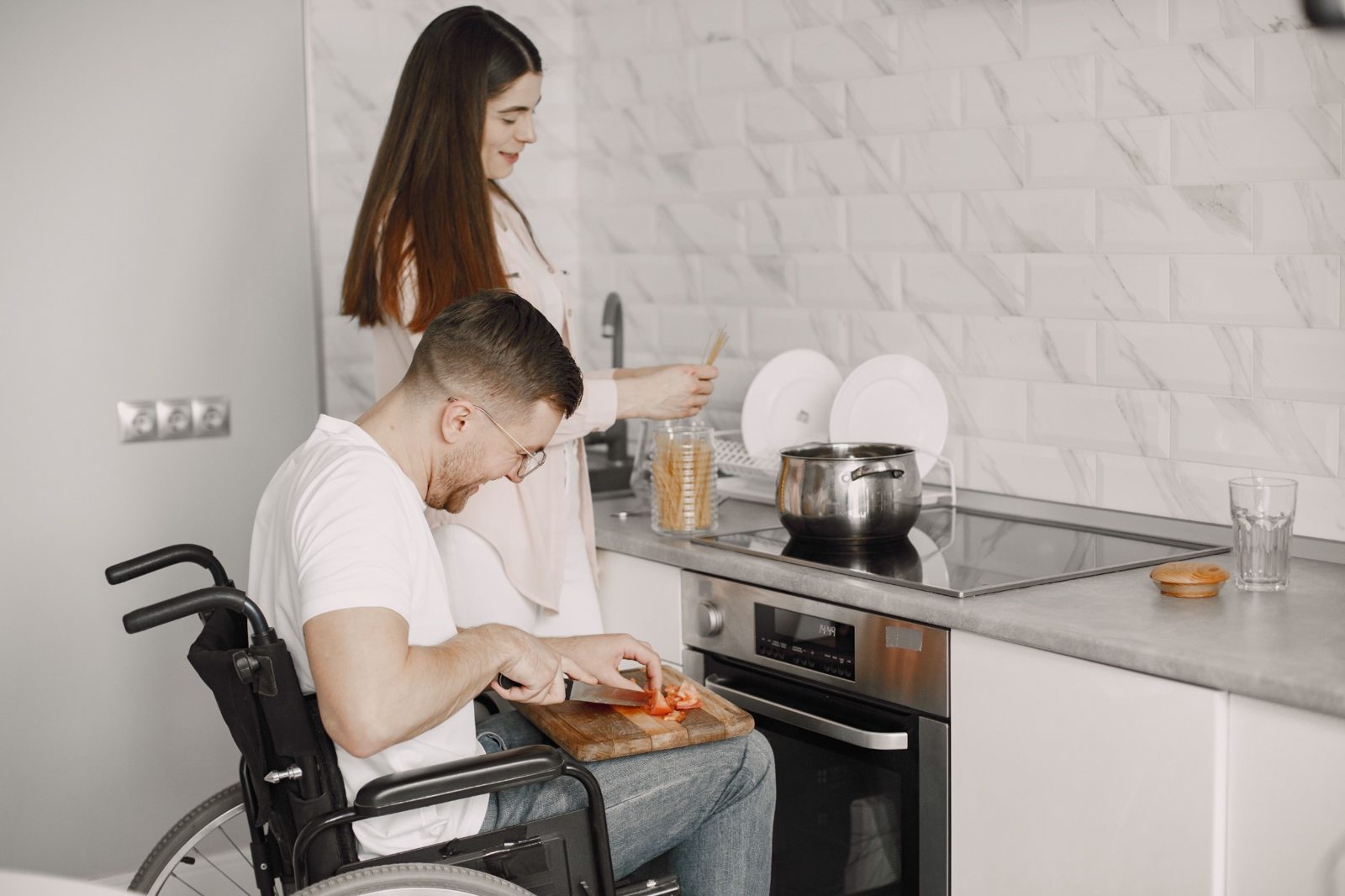 A man in a wheelchair chops tomatoes on a board on his lap while a housemate cooks pasta at the stove.